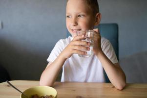 un' carino ragazzo bevande acqua e mangia un' asciutto prima colazione a casa e sorrisi foto