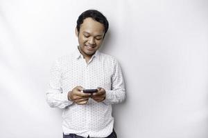 eccitato asiatico uomo indossare bianca camicia sorridente mentre Tenere il suo Telefono, isolato di bianca sfondo foto