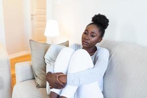 triste africano americano donna con depressione seduta su divano. solitario stressato irritato giovane nero ragazza pensiero di psicologico problema pensiero rimpiangere di sbaglio foto