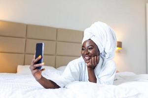 sorridente africano giovane donna dire bugie su letto nel accappatoio con sua mobile Telefono assunzione un' autoscatto. africano americano donna rilassante su il letto dopo bagno e guardare a il Telefono telecamera assunzione un' autoscatto foto