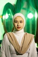 un' musulmano donna con un' bianca foulard e bianca Abiti è seduta nel il mezzo di un' verde camera senza trucco foto