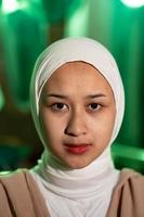 un' musulmano donna con un' bianca foulard e bianca Abiti è seduta nel il mezzo di un' verde camera senza trucco foto