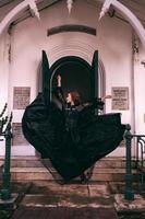un elegante strega colpi di frusta sua tutti nero vestito nel il aria nel davanti di il cimitero cancelli dopo il Halloween Festival inizia foto