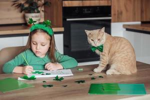 un' poco ragazza con un' bendare su sua testa disegna e tagli verde quadrifogli per st. Patrick giorno a un' tavolo a casa nel il cucina, Il prossimo per sua è sua bellissimo gatto con un' verde arco cravatta in giro il suo collo foto