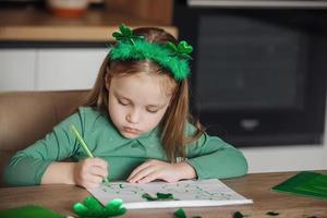 un' poco ragazza con un' trifoglio fascia per capelli disegna e tagli verde quadrifogli per st. Patrick giorno a sua tavolo a casa foto