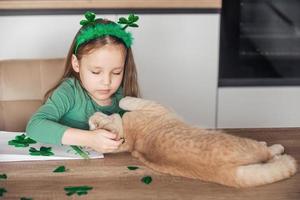 un' poco ragazza con un' bordo decorato con trifoglio disegna e tagli verde quadrifogli per st. Patrick giorno a il tavolo a casa nel il cucina, Il prossimo per sua è sua bellissimo gatto foto