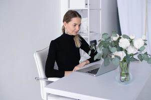 il ragazza lavori nel un' bianca ufficio con un' il computer portatile e fiori su il tavolo foto