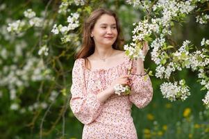 un' ragazza nel un' rosa vestito sta vicino un' bianca albero con fiori sembra lontano e sorrisi foto