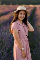 un' allegro giovane ragazza nel un' rosa vestito e un' cannuccia cappello nel sua mani sta tra lavanda cespugli. tramonto. foto