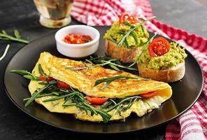 prima colazione. frittata con pomodori, formaggio, verde rucola e toast con avocado crema su nero piatto. frittata - italiano frittata. foto