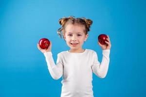 carino poco ragazza con riccioli nel un' a strisce vestito è Tenere un' grande rosso Mela nel sua mani. blu sfondo. utile prodotti per bambini. salutare merenda. spazio per testo. alto qualità foto