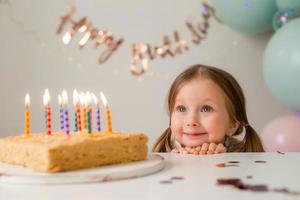 carino poco ragazza colpi su candele su un' compleanno torta a casa contro un' fondale di palloncini. del bambino compleanno foto