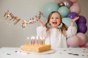 carino poco ragazza colpi su candele su un' compleanno torta a casa contro un' fondale di palloncini. del bambino compleanno foto