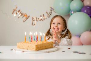 carino poco ragazza colpi su candele su un' compleanno torta a casa contro un' fondale di palloncini. del bambino compleanno foto