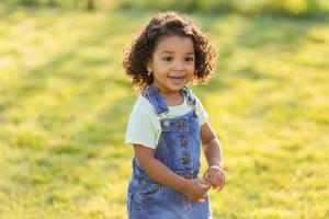 ritratto poco contento giocoso bambino piccolo bruno ragazza nel un' denim prendisole in piedi nel il giardino su un' soleggiato giorno. a piedi nel il fresco aria. concetto di un' contento infanzia. spazio per testo. alto qualità foto