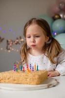 carino poco ragazza colpi su candele su un' compleanno torta a casa contro un' fondale di palloncini. del bambino compleanno foto