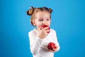 carino poco ragazza con riccioli nel un' a strisce vestito è Tenere un' grande rosso Mela nel sua mani. blu sfondo. utile prodotti per bambini. salutare merenda. spazio per testo. alto qualità foto