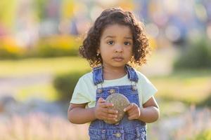 ritratto poco contento giocoso bambino piccolo bruno ragazza nel un' denim prendisole in piedi nel il giardino su un' soleggiato giorno. a piedi nel il fresco aria. concetto di un' contento infanzia. spazio per testo. alto qualità foto