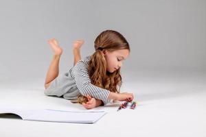 poco ragazza nel un' a strisce vestito disegna nel un album dire bugie su il pavimento. bambino sviluppo. stile di vita. prescolastico e scuola formazione scolastica. spazio per testo. alto qualità foto