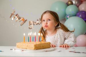 carino poco ragazza colpi su candele su un' compleanno torta a casa contro un' fondale di palloncini. del bambino compleanno foto