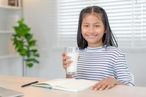 bambini donne bevanda latte per nutrire il corpo e nutrire il cervello. asiatico giovane poco ragazza imparare a casa. ragazza contento bevanda latte e leggere un' libro per esame, scuola a casa. formazione scolastica, vitamine, sviluppo foto