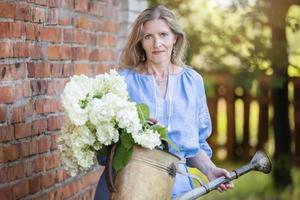 un' bellissimo anziano donna nel un ricamato camicia detiene un' retrò irrigazione può con fiori. il concetto di floricoltura e giardinaggio come un' passatempo. foto