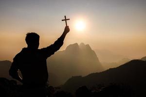 silhouette di giovane uomo preghiere e Tenere cristiano attraversare per adorazione Dio a tramonto sfondo. cristiano religione concetto sfondo. foto