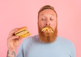 riflessivo uomo con barba e tatuaggi mangia un' sandwitch con Hamburger e patate foto