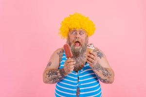 Grasso uomo con barba e parrucca mangia un' ghiacciolo e un gelato foto