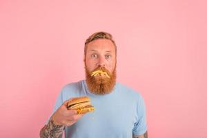 Affamato uomo con barba e tatuaggi mangia un' sandwitch con Hamburger e patate foto