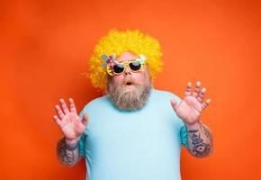 Grasso stupito uomo con barba, tatuaggi e occhiali da sole ha divertimento con il giallo parrucca foto