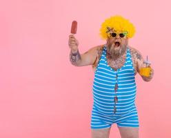 Grasso sorpreso uomo con barba e parrucca mangia un' ghiacciolo e bevande un' succo frutta foto