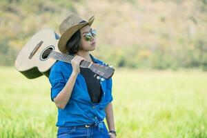 donna indossare cappello e trasportare sua chitarra nel erba campo foto