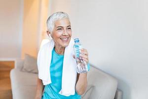 anziano donna seduta su il divano, esausto dopo il quotidiano training.senior donna assunzione un' rompere mentre esercizio a casa. atletico maturo donna Tenere bottiglia di acqua, avere un' asciugamano in giro sua collo foto