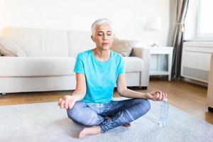 anziano donna praticante yoga, seduta nel metà loto esercizio, ardha padmasana posa, Lavorando fuori, indossare abbigliamento sportivo, meditazione sessione, interno pieno lunghezza, casa interno foto