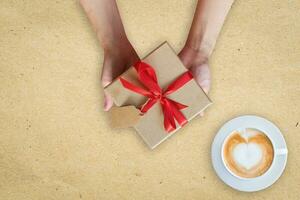 vicino su mano donna Tenere regalo scatola su Marrone carta con caffè tazza. foto