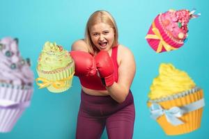 Grasso ragazza nel fitness suite fa boxe e combattimento contro cupcakes. ciano sfondo foto