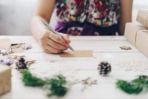 vicino su di mani donna scrittura vuoto Lista dei desideri e Natale carta su di legno tavolo con natale decorazione. foto