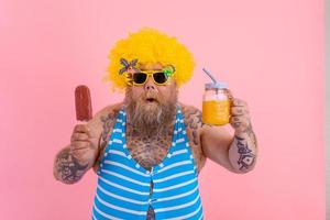 Grasso sorpreso uomo con barba e parrucca mangia un' ghiacciolo e bevande un' succo frutta foto