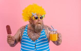 Grasso pensieroso uomo con barba e parrucca mangia un' ghiacciolo e bevande un' succo frutta foto