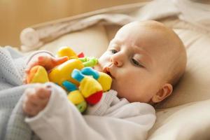 un' piccolo bambino bugie e giochi con un' giocattolo. bambino con un' giocattolo nel il suo bocca foto