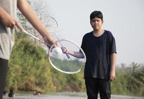 asiatico ragazzi hold badminton volano e racchetta, in piedi e giocando accanto il fiume banca nel loro Locale fiume durante loro fine settimana vacanza, morbido e selettivo messa a fuoco su davanti ragazzo nel bianca camicia. foto