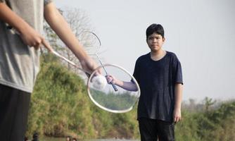 asiatico ragazzi hold badminton volano e racchetta, in piedi e giocando accanto il fiume banca nel loro Locale fiume durante loro fine settimana vacanza, morbido e selettivo messa a fuoco su davanti ragazzo nel bianca camicia. foto