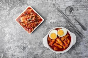 superiore Visualizza tteokbokki o speziato riso torte con bollito uova, kimchi lato piatto e inossidabile acciaio cucchiaio e bastoncini su il grigio calcestruzzo tavolo, coreano strada cibo. foto