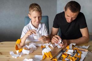 un' carino ragazzo è seduta con il suo papà a il tavolo e raccolta origami. colla il parti foto