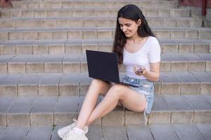 il ragazza è seduta su il scale, il computer portatile su sua giro, guardare in il distanza foto