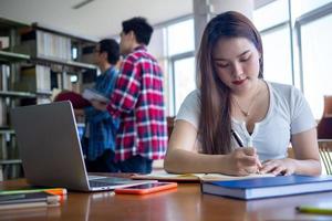 giovane asiatico studenti nel casuale indossare fare compiti a casa e uso un' tecnologia il computer portatile nel il Università biblioteca. il studenti siamo ricerca per conoscenza e siamo preparazione per il esame. foto