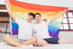 contento asiatico lgbt coppia, seduta su il divano Tenere e agitando arcobaleno lgbt orgoglio bandiera insieme nel il Camera da letto a casa. diversità di lgbt relazioni. un' gay coppia concetto. amorevole gay coppia. foto