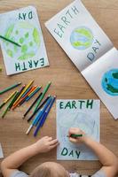 un' bambino a casa a il tavolo disegna il pianeta terra con un' mondo carta geografica con multicolore matite e pennarello penne su bianca carta. pace e terra giorno concetto. foto