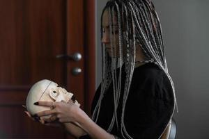 un' giovane luminosa ragazza nel buio Abiti, con dreadlocks su sua testa, detiene un' cranio nel sua mani. generazione z, Gotico stile foto